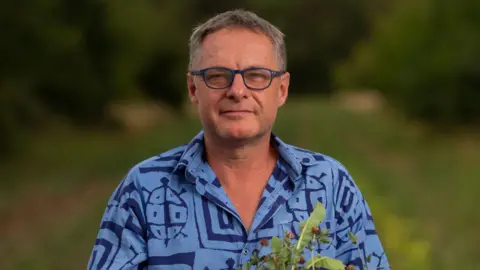 Contributed David Wolfe is pictured smiling to camera while standing in a field. He has short grey hair and is wearing a blue shirt with patterns on it. He is wearing dark coloured glasses and he is holding a plant.