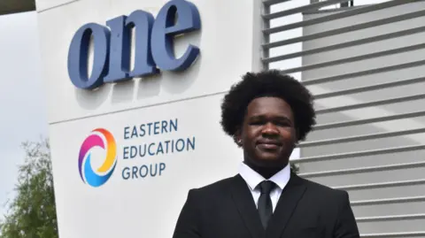 John Nice DJ Mudziviri wearing a black suit jacket and tie over a white shirt. He is standing in front of Suffolk One Sixth Form College, the logo for which can be seen to the left of the image. He is looking and smiling directly into the camera