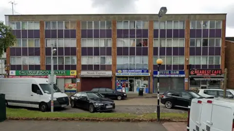 Google Street view of a three-storey brick building with a row of ground-floor shops, including “Dallow Food Centre”, “Snack Express”, a glass and glazing services shop and “Nearest Cafe”. Several cars and two white vans are parked along the roadside in front. There is a grass verge with a streetlight and a beacon on top of a black and white post.