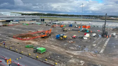 Nicola Haseler/BBC The site of the former car park. There are many construction vehicles and workers. The site is located next to a small runway and is mostly covered in mud.