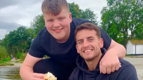 Josh McDonald wears a blue hoody and smiles at the camera. His younger brother Max wears a blue T-shirt and crouches beside Josh and puts his arm around him.