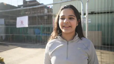 A young girl with dark hair, wears a grey Nike jacket, and smiles in the sunshine 