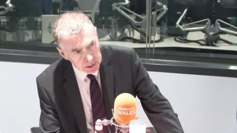 Adam Price in a radio studio looking out of shot at his interviewer in front of an orange microphone with BBC Radio Wales branding white letters.