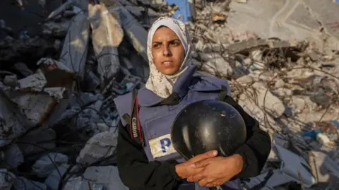 AP Freelance journalist Mariam Dagga, wearing a blue Press vest, a beige headscarf and holding a black helmet with clutched to her body with both hands, shown in front of mangled concrete and iron from the rubble of a building.