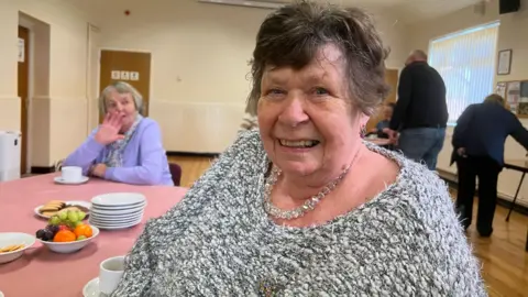 Shirley Coombes. She is sat at a table. She has short dark hair and is wearing a grey jumper with a necklace. She is looking directly at the camera and smiling. 