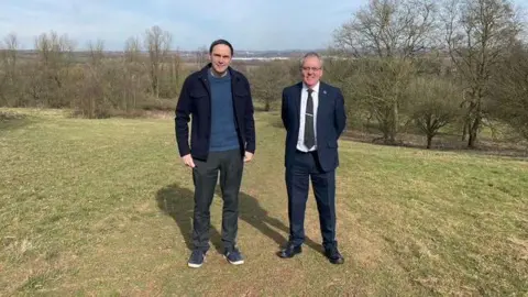 Two men standing in the middle of a field in the countryside. Trees are visible in the background.