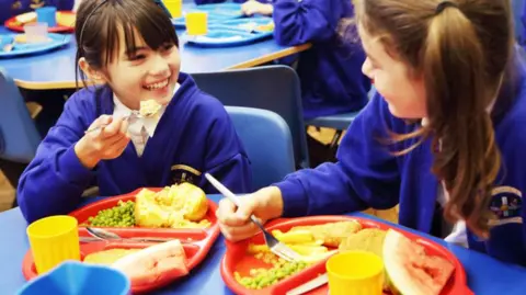 Getty Images Two school girls in blue uniforms smile to each other as they eat school dinners from a plastic tray