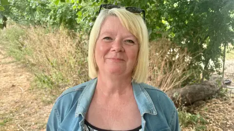 Leanne Brown/BBC A middle-aged woman with blonde hair cut into a bob stands in front of greenery. She is looking into the camera with a closed-mouth smile, wearing a blue denim jacket over a black and white patterned top, and sunglasses placed on her head.