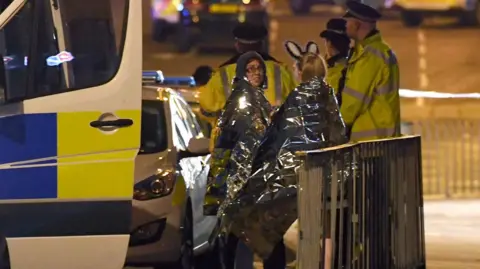 Getty Images Concert goers wrapped up in silver foil survival blankets wait to be picked up at the scene outside the Manchester Arena  