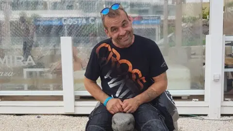 Family Carl Dyche sitting on a wall wearing a T-shirt with sunglasses on his head, looking happy.