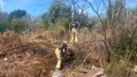 Photograph of two firefighters tackling a blaze at an illegal waste dump in Ashton-upon-Mersey. The image shows a them using a hose.