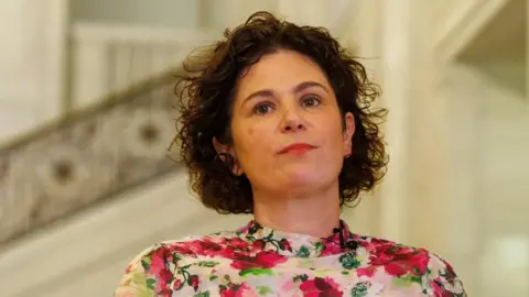 PA Media A close-up image of Claire Hanna in front of a blurry background. She hs curly brown hair and is wearing a white top with pink and green flowers.