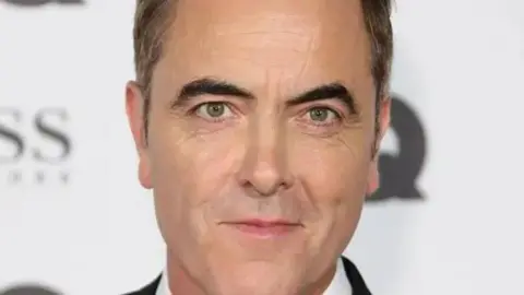 Getty A man with black hair and black eyebrows is attending a red carpet event in a tuxedo. A number of blurred sponsors can be seen in the background wall.