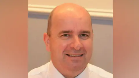 Halton Borough Council A middle-aged man with very short hair wearing a white shirt smiles at the camera. 