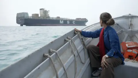 A woman sits in a small grey boat looking at a large cargo ship on the sea with the letters MSC written in white on its side. Her long, dark hair is tied up and she is wearing a blue top. There are some orange life jackets behind her. 