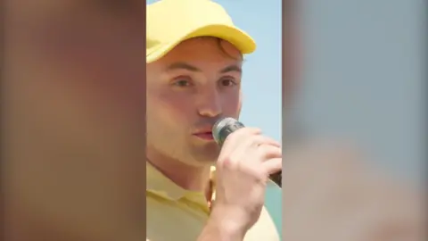 A man speaks into a mic. He wears a yellow baseball cap and T-shirt.