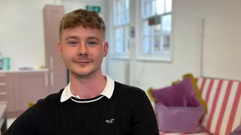 Ben Schofield/BBC Jay Virgo looking directly down the camera. He is wearing a black pullover top with a white collared t-shirt underneath. He is sitting down and smiling. He has light brown hair, cut short at the sides, blue eyes and has small earrings in each ear. He is seated inside a room with white walls, two large sash-style windows and some light purple-coloured cupboards against one wall. On the right of the image is a sofa with a red and white striped back and two purple cushions.