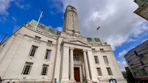 A picture of the town hall in Luton, which is the headquarters of Luton Council. It is a grey building with four flours, and has a clock tower on top of it. The sky is a mixture of blue but also with some light grey clouds behind the building.