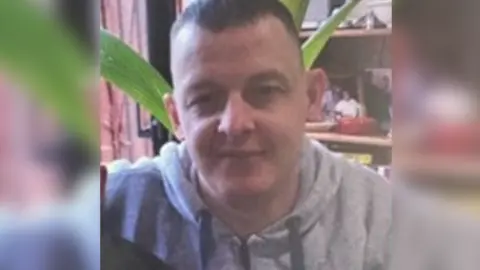 A family handout photo of Paul Rowe. He is smiling into the camera. He has short brown hair and is wearing a grey hoodie.
