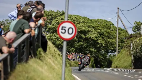 IOM TT Crowds are leaning on a fence on the left on a verge overlooking a road to the right below them. There is a motorbike racer travelling away and there are trees in the background. In the front of the shot is a pole with a 50mph speed limit sign on it.