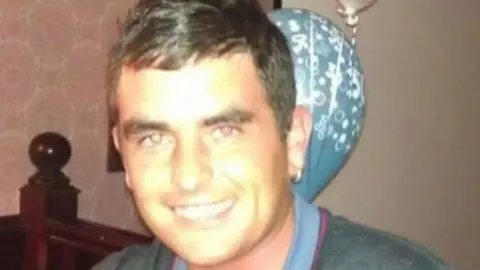 Craig Hamilton, a smiling man, with short dark hair combed forward. He has a gold hooped earring in his left ear.