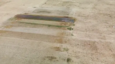 A drone shot of Plymouth Cricket Club's playing surface dry and parched. The wicket has a singular hose pipe on it and is very dark coloured.