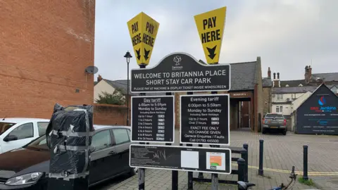A car park sign which reads "Welcome to Britannia Place short stay car park." The larger sign consists of four separate black signs detailing day and evening tariffs. In addition there are two large upturned yellow pyramids on top with the words "pay here" written on them. A couple of parked cars can be seen on the left.