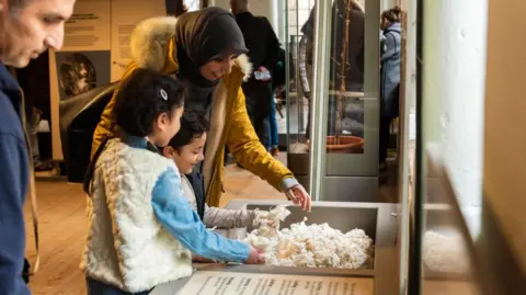 National Trust Images A young girl and boy play with material which is part of an exhibition at Quarry Bank as their mother looks on. The girl who has her back to the camera has her hair in a plait. The boy is smiling as he plays.  