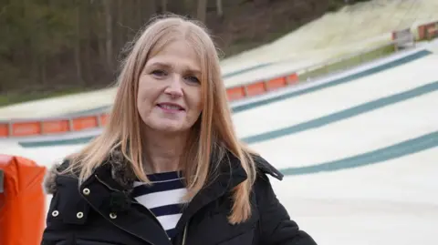 Shaun Whitmore/BBC Beki Cozens is standing outside in front of a dry ski slope. She is looking directly at the camera and is smiling. She has blonde hair and is wearing a black outdoor coat and a striped black and white T-shirt.