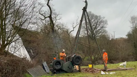 Two workmen wearing hard hats and orange high-visibility jackets with a huge percussive borehole - a metal structure at least 20ft high, with wheels at the base. There's smoke coming out of the exhaust - it's almost like an old fashioned traction engine. Behind the borehole you can see some of the white sheets covering the damaged bridge. It's on the edge of a field, with bare trees surrounding it. The sky is grey and overcast. 