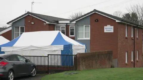 A two storey block of flats with a forensic tent over the garden area outside. A sign says Gerards Court.