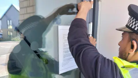 A police officer, putting a white notice on a glass door. One arm is raised and the other is on the glass. He is wearing a police hat, blue top and high-vis jacket. He has a beard, a small earring and short dark hair. 