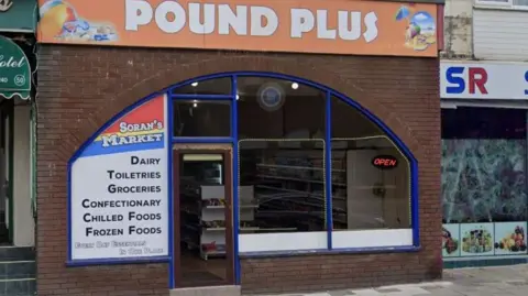 Google A food shop on Coronation Street in Blackpool with an orange sign above it saying Pound Plus and a blue lined window saying Soran's Market - selling Dairy, Toiletries, Groceries - a neon red sign says Open in the corner