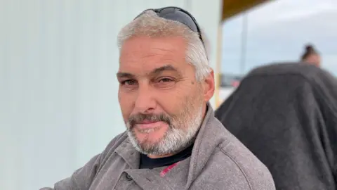 Henry Godfrey Evans/BBC Christopher looking at the camera. He has a beard and is wearing a grey jumper.