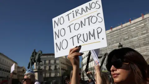 Reuters A dark-haired woman wearing sunglasses holds a sign that reads: "No tyrants, no fools, no Trump"