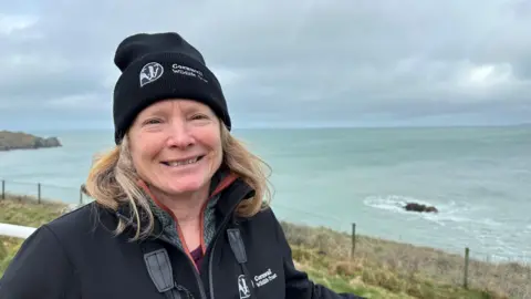 A woman stood on a the coast wearing a black beanie hat and fleece which both say 'Cornwall Wildlife Trust' on them. It is a misty day and the woman is smiling