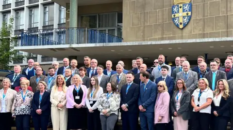 Lots of people in smart work clothes posing for a photo in rows outside Durham County Hall. Above them is the council logo on the walls of the building. The logo is blue and has a yellow cross. 