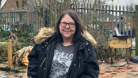 A woman with dark brown hair and glasses is wearing a black hooded coat and a dark grey tshirt with star and moon design on the front. She has a slight smile and is looking directly at the camera.