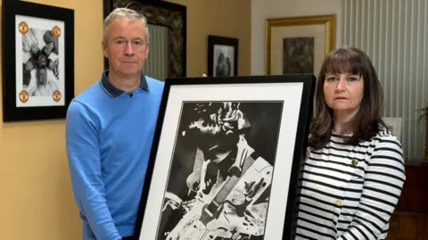 Peter Dolan and his wife Niamh Dolan standing together and looking at the camera. Between them they are holding a larger black and white photo of their late son Enda playing guitar.