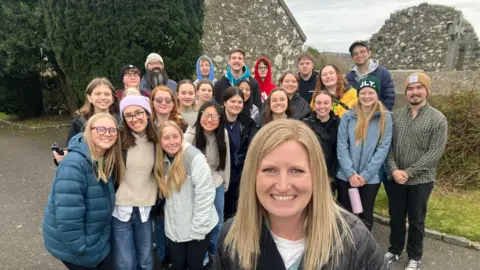 Brittany Shoemake poses for a photo with a group of 23 young US tourists at Saul Church in County Down.   Brittany has shoulder-length blonde hair and is wearing a white t-shirt under a black jacket.  Some of the youg people are weaing padded jackets and woolen hats. 