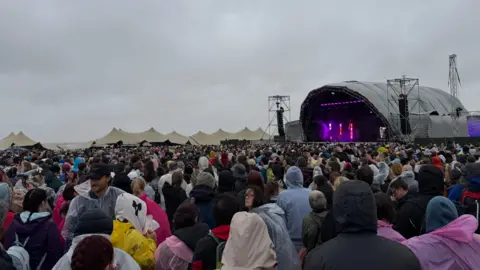 Cloudy, rainy skies above a purple-lit stage, which tent roofs visible in the distance. There is a large crowd mostly dressed in waterproofs and ponchos.