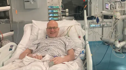 GRAHAM MCMILLAN Graham McMillan is lying in a hospital bed wearing a hospital gown and smiling at the camera while propped up by pillows. There is medical equipment and screens next to his hospital bed, including several wires attached to him.