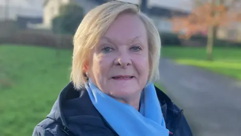 A woman with short-cut blonde hair smiles as she looks at the camera. She is standing in a park with trees, grass and a walkway behind her. The woman is wearing a navy rain coat and a bright, light blue scarf around her neck. 