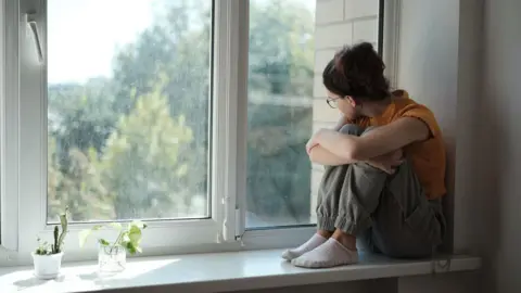 A teenage girl sitting on a window sill looking outside. She is wearing cargo pants and an orange top. She has brown hair and is wearing glasses but is facing away from the camera. Two small plants are in pots on the left side of the window sill.