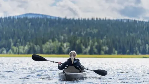 Getty Images A woman, with blonde hair, in a small boat with a paddle canoe, on a stretch of water, with trees in the background.