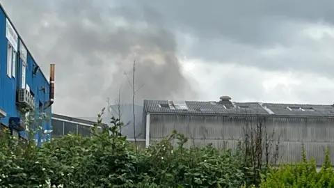 Emma Baugh/BBC Grey warehouse building to the right and blue two-storey factory to the left. There are trees in the foreground. Grey smoke is rising from the area behind the warehouse.