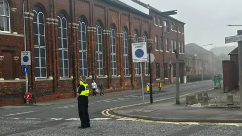 Amber Gash/BBC A police officer wearing black trousers and yellow high visibility vest stands in the centre of a road. A large brick building is visible in the background which is covered in hazy grey smoke.
