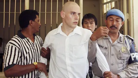 Getty Images Indonesian police escort Matthew Norman and Si Yi Chen, who are both young men, to Denpasar District Courtroom for their sentencing trial. Norman wears a white shirt in the foreground. His arms are being held by Indonesian prison guards. A barred cell is open in the background.