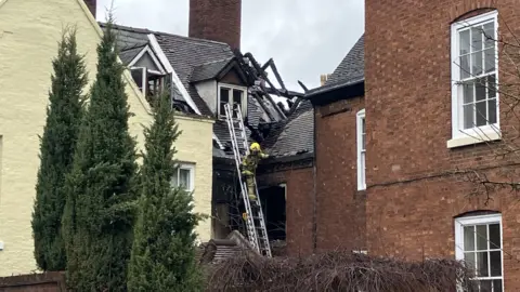 BBC A firefighter with a yellow helmet on a ladder against a property. The roof beams are exposed after a fire and they are charred.