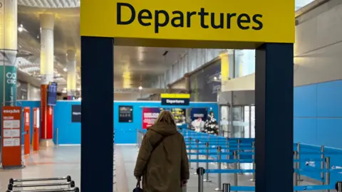 A woman walking through departures area at Jersey Airport. A Christmas tree can be seen in the foreground at the second entry.